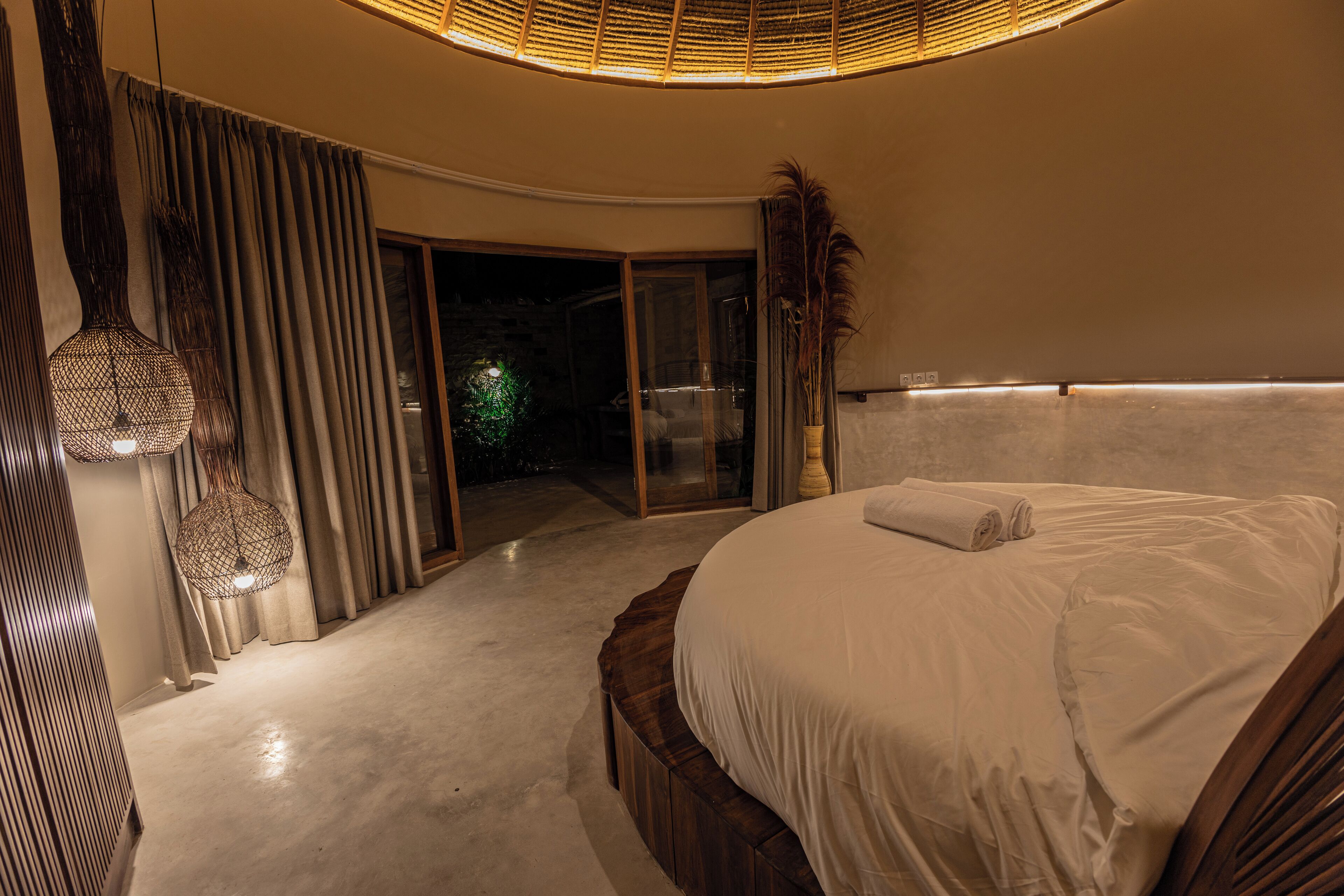 A bedroom with a large bed, a wooden headboard, a glass door leading to a balcony, and a hanging light fixture.