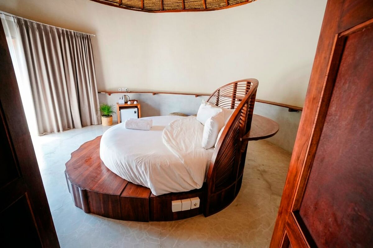 Una cama redonda con estructura de madera, una mesita de noche, una lámpara y una planta en maceta en una habitación con techo curvo y un gran ventanal con cortinas.
