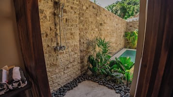 Un espace de douche aux murs en pierre, doté d’une pomme de douche intégrée, entouré d’une bordure de galets et d’une végétation tropicale luxuriante.