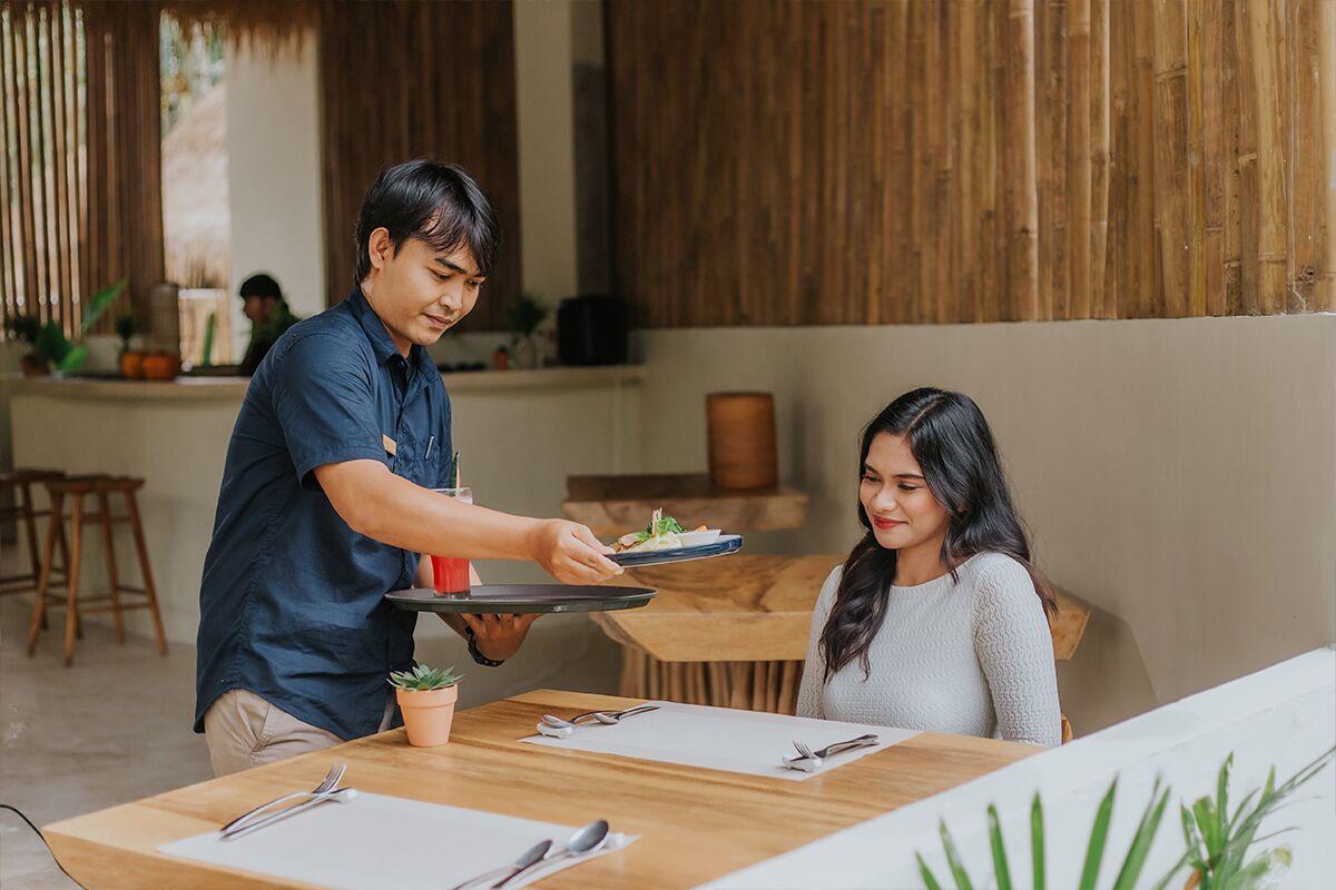 A person serving food to another person at a table in a room with wooden walls and a plant on the table.