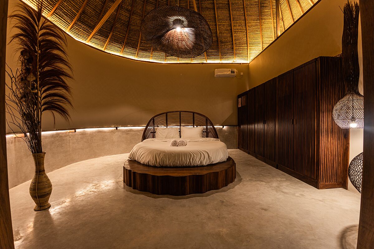Una cama redonda con estructura de madera, un jarrón grande con ramas y una lámpara de techo con textura.