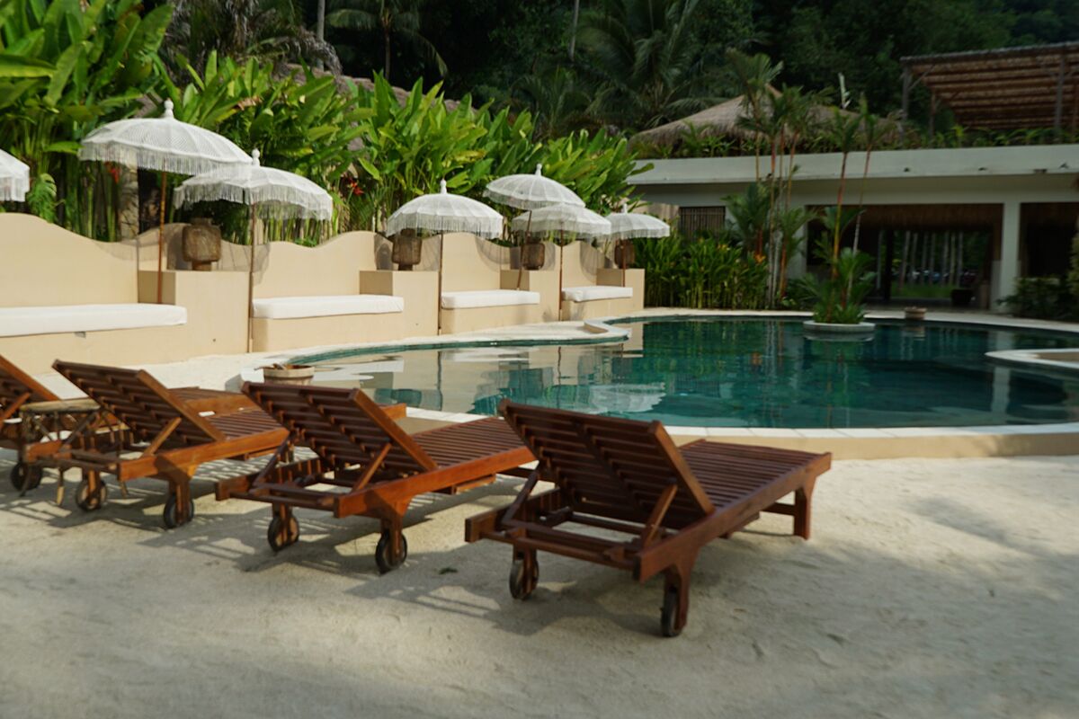 Uma área à beira da piscina com espreguiçadeiras e guarda-sóis, cercada por vegetação.