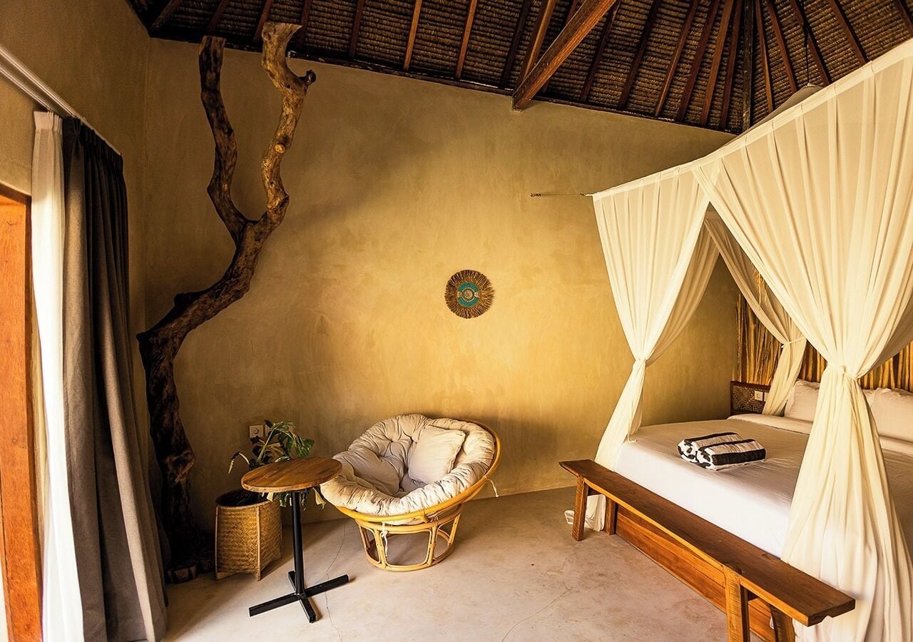 A room with a bed, a chair, a small table, a plant, and a unique wall decor.