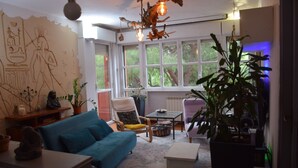 Ein Wohnzimmer mit einem blauen Sofa, einem Holzstuhl, einem Couchtisch und einer großen Topfpflanze. Ein Fenster bietet Blick auf Bäume, und an der Wand hängt ägyptische Kunst.