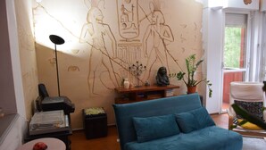 Ein Wohnzimmer mit einem blauen Sofa, einer Wand mit ägyptischen Hieroglyphen, einem Drucker, einem kleinen Tisch mit einem roten Gegenstand und einer Topfpflanze.