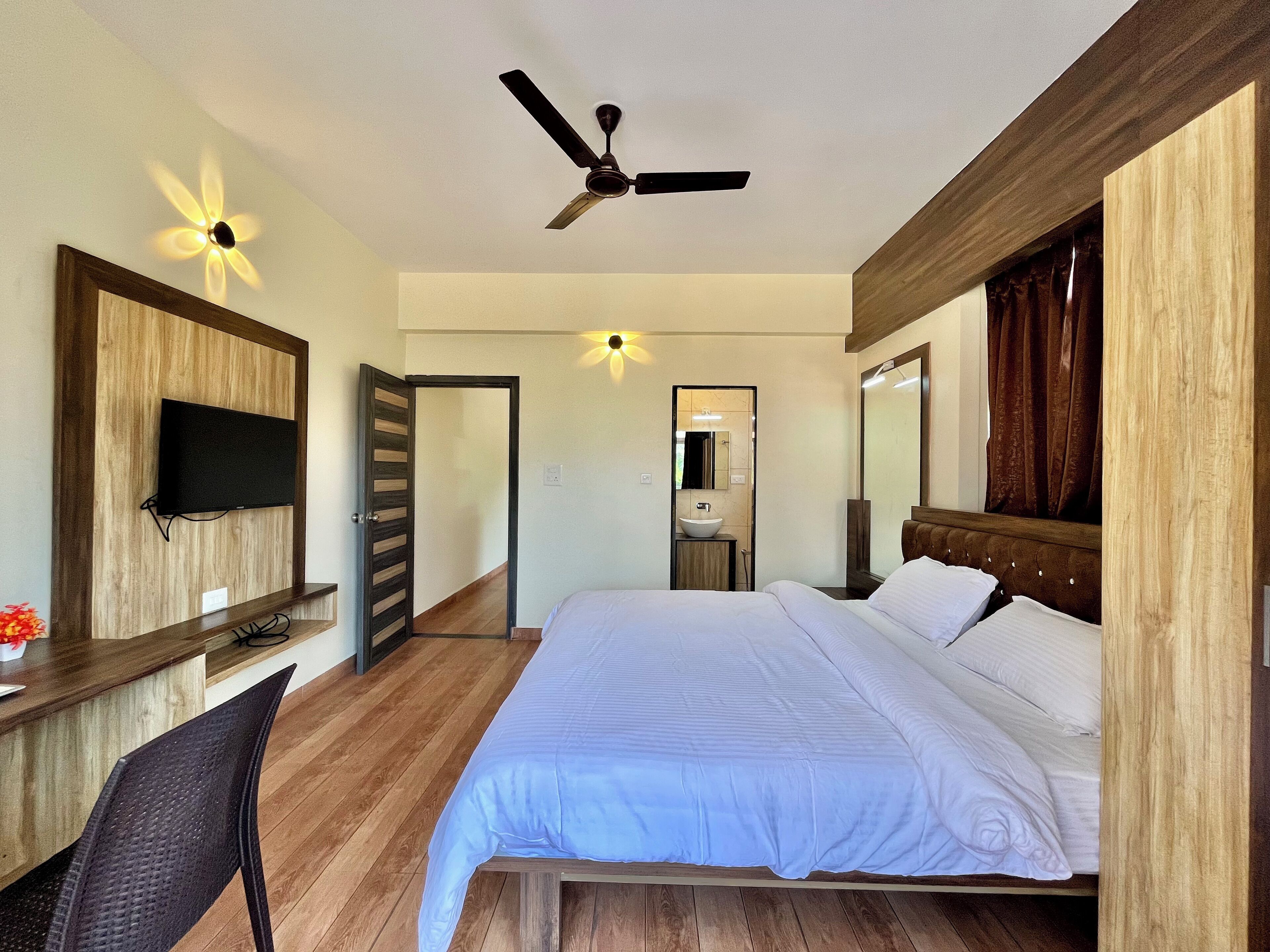 Una camera d'albergo con un letto, una scrivania con televisore, una ventola a soffitto e un bagno visibile attraverso una porta aperta.