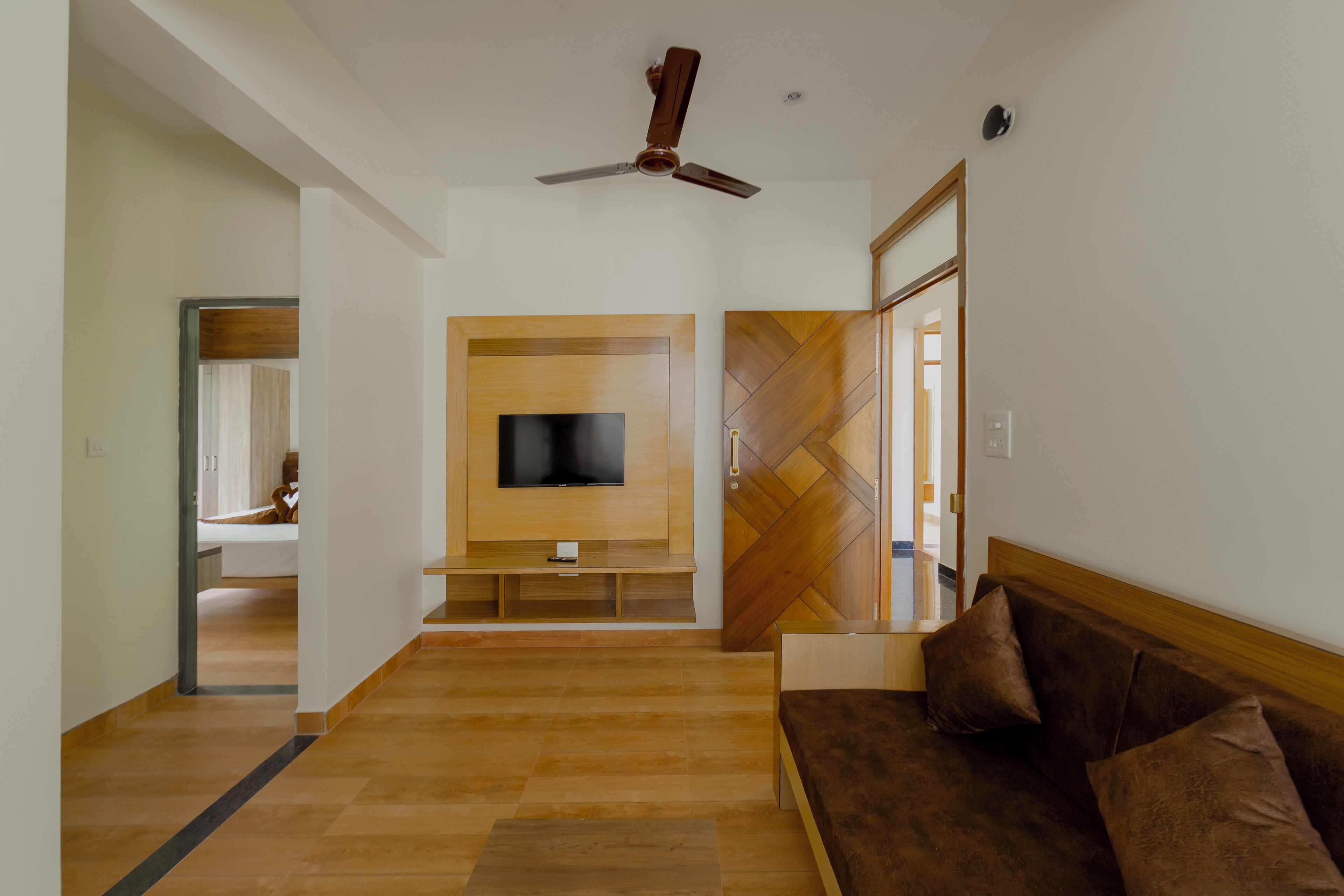 A modern living room with a wooden floor, a brown sofa, a television on a wooden stand, and a ceiling fan.