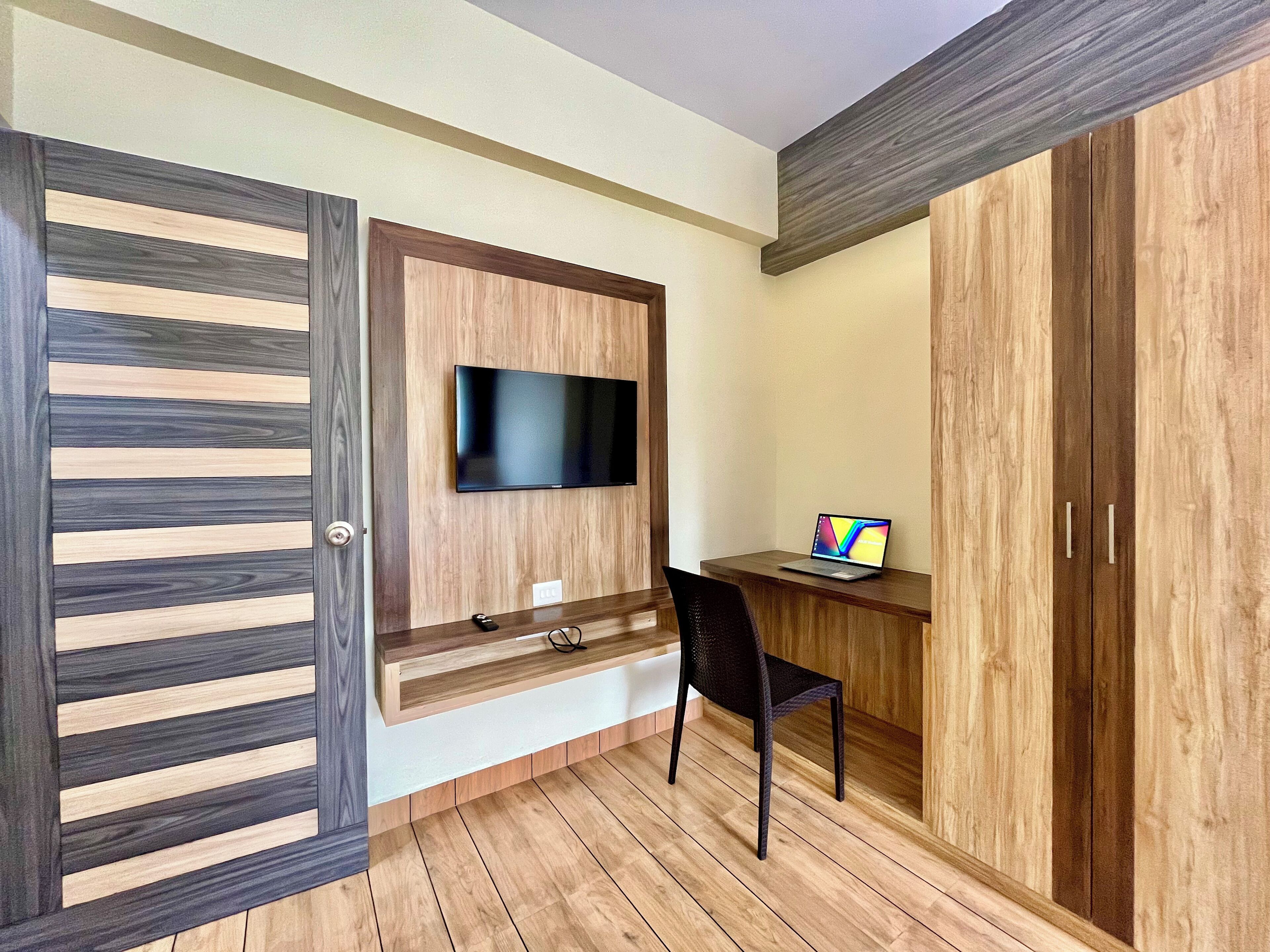 Una habitación moderna con suelo de madera, un televisor montado en un panel de madera, un escritorio con una computadora portátil y una silla.