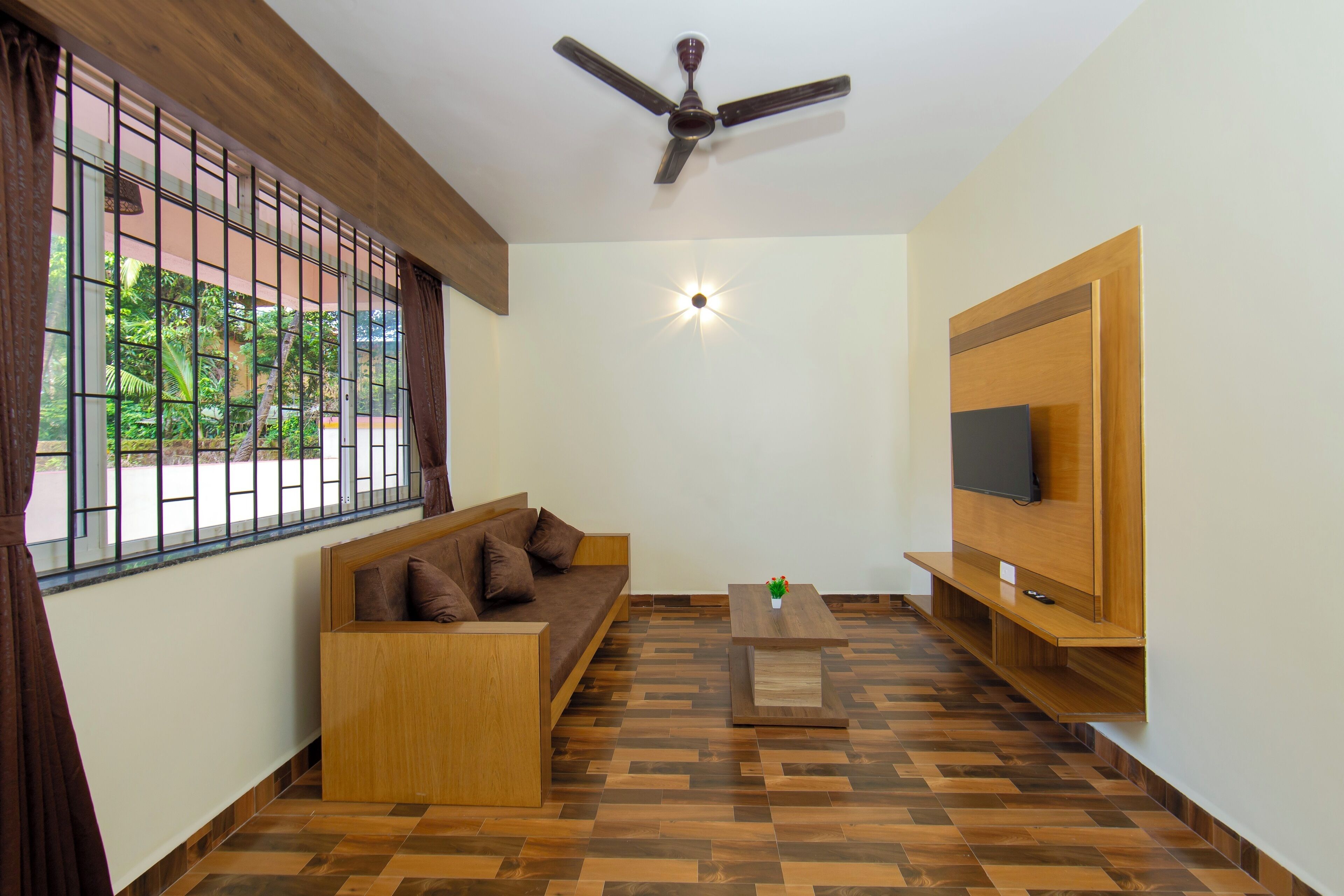 A room with a wooden sofa, a TV mounted on the wall, a ceiling fan, and a window with a view of greenery.
