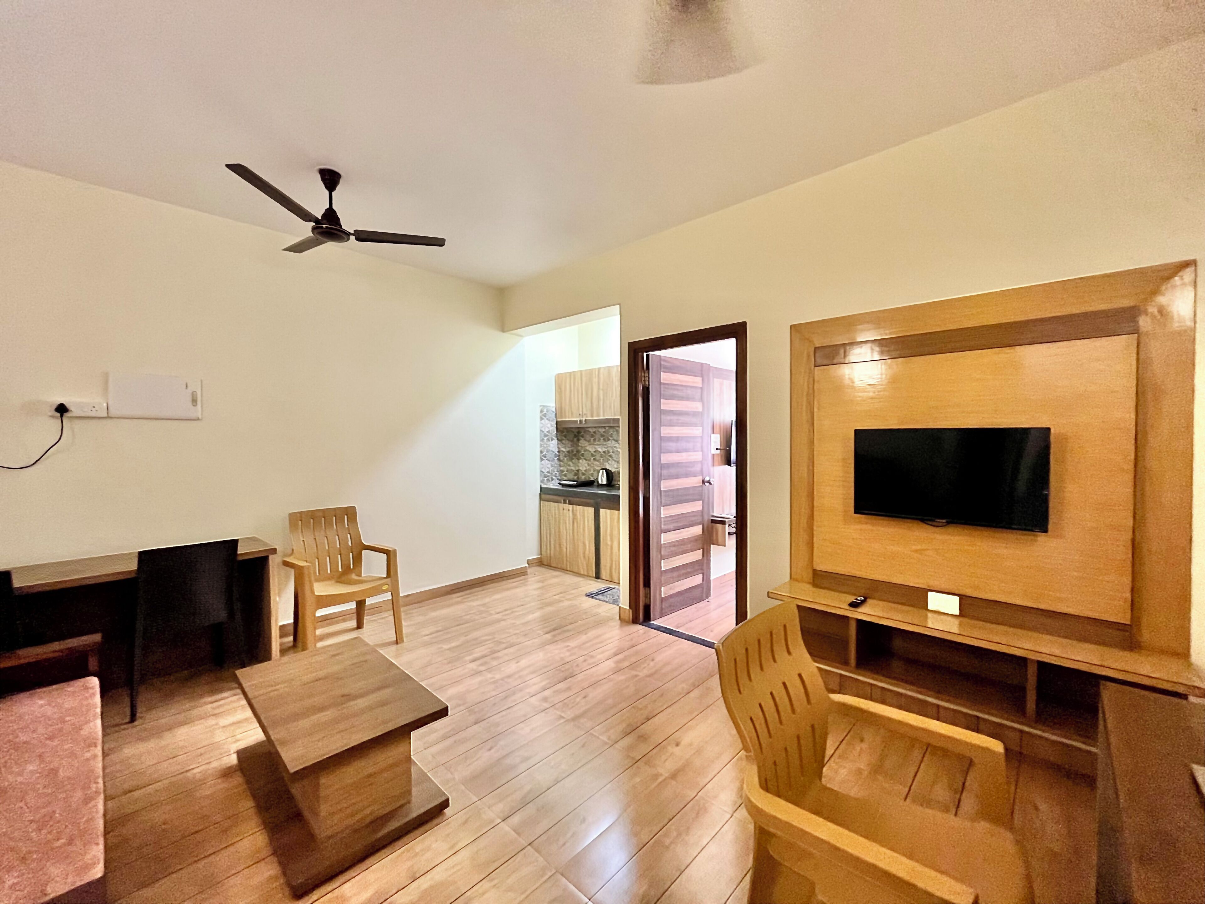 A living room with a TV mounted on the wall, wooden furniture, a ceiling fan, and a kitchen visible through an open door.