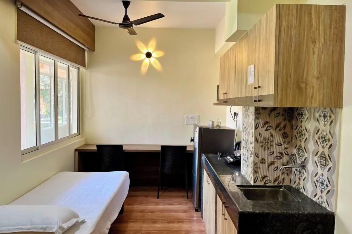 Una habitación compacta con cama, escritorio, silla, ventilador de techo, lámpara de pared y zona de cocina con lavabo y armarios.