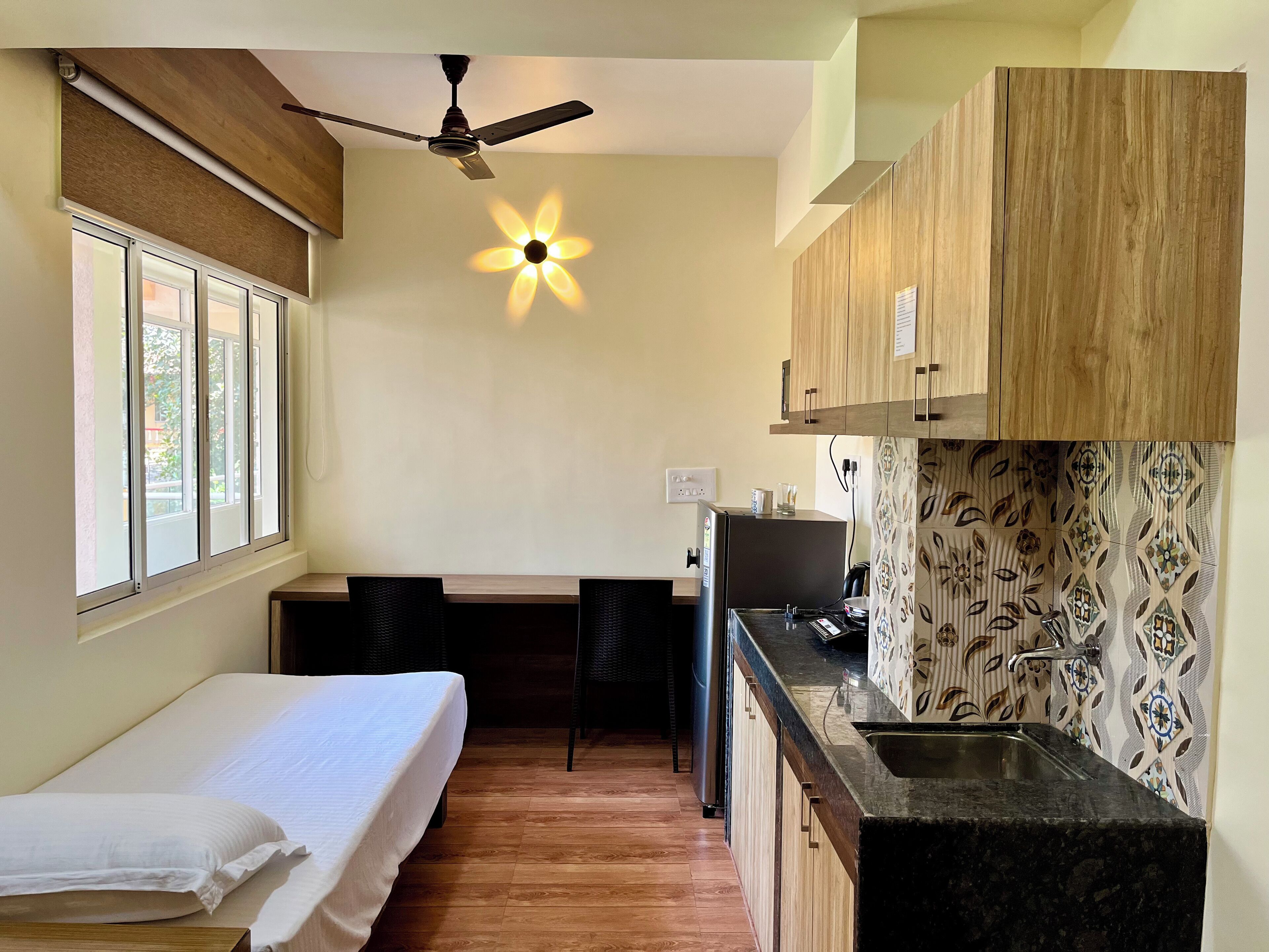 A compact room with a bed, a small kitchenette, a ceiling fan, and a window.