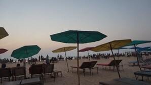 Una playa con hileras de sillas y sombrillas, arena y gente a lo lejos.