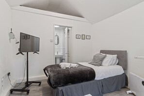 A bedroom with a bed, a TV on a stand, a bathroom visible through an open door, and a small shelf with framed pictures. - Studio Cottage W/Shared Garden - 3Min to Bus stop (Manchester)