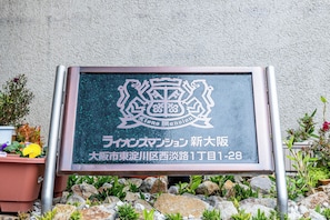 Um letreiro com um brasão e texto indicando 'Lions Mansion' em japonês, localizado em '1-28, 1º beco, 1º quarteirão, Bairro Naka, Cidade de Nagoya, Prefeitura de Aichi'.