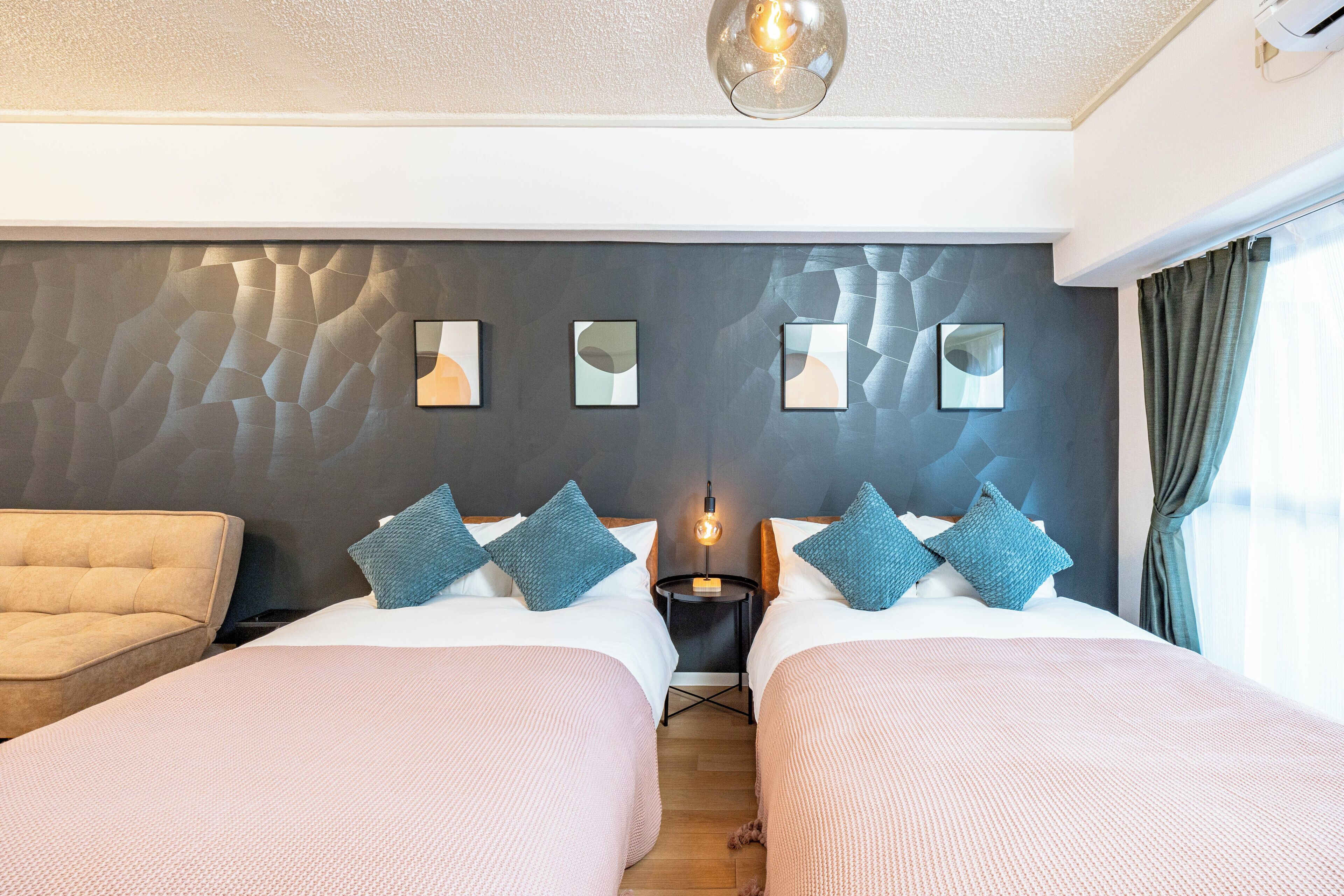 Ett hotellrum med två sängar, en soffa, en väggmonterad lampa och ett fönster med gardiner.