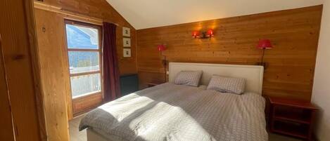 A bedroom with a wooden ceiling, a bed with patterned bedding, two bedside tables with lamps, and a window with curtains.