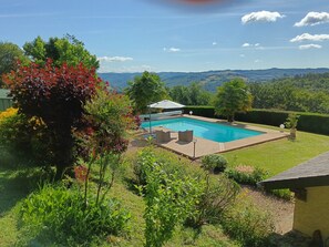 A rectangular swimming pool with a wooden deck, surrounded by lush greenery and a view of distant hills. - Welcome to our Gîte   (Naves)