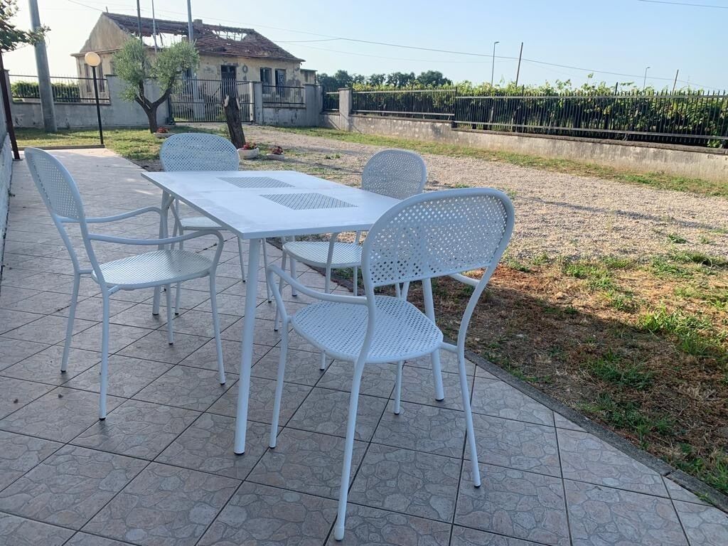 Een terras met een witte tafel en vier stoelen, omringd door een betegelde vloer en een gazon met een hek op de achtergrond.
