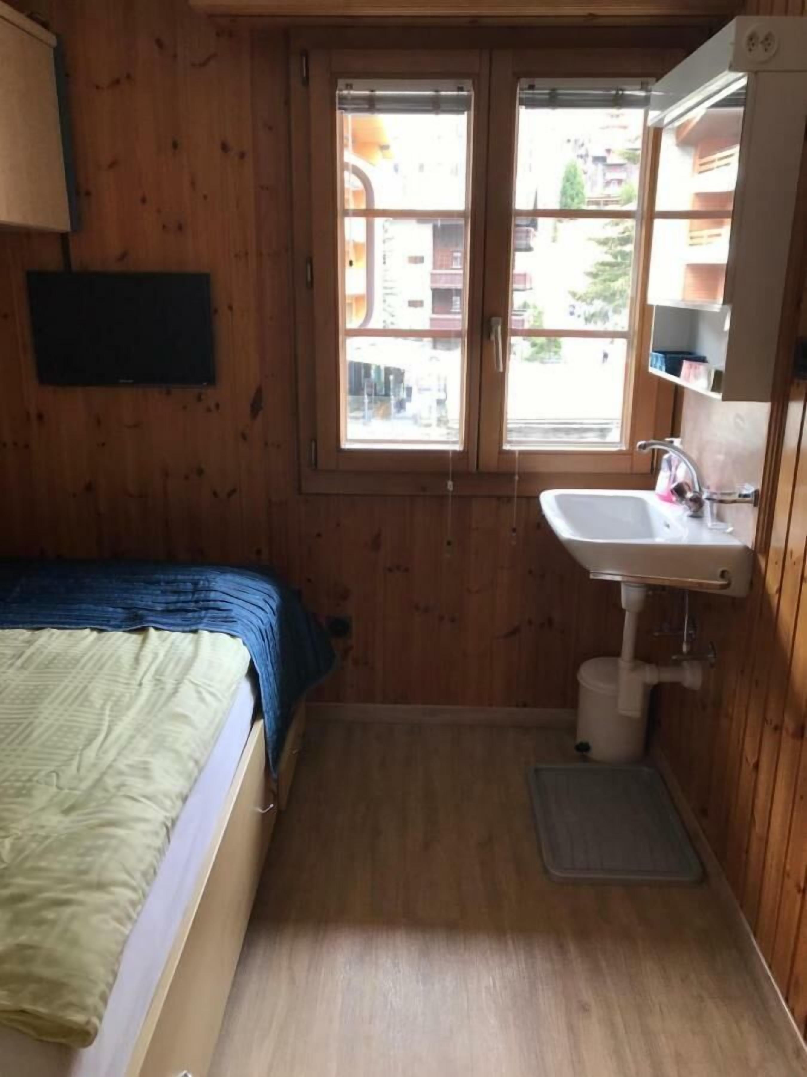 A small, wooden-paneled room with a bed, a window, a sink, and a mirror.