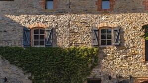 Un bâtiment en pierre avec des fenêtres en arc et des volets, une partie du mur recouverte de lierre, et une porte en bas.