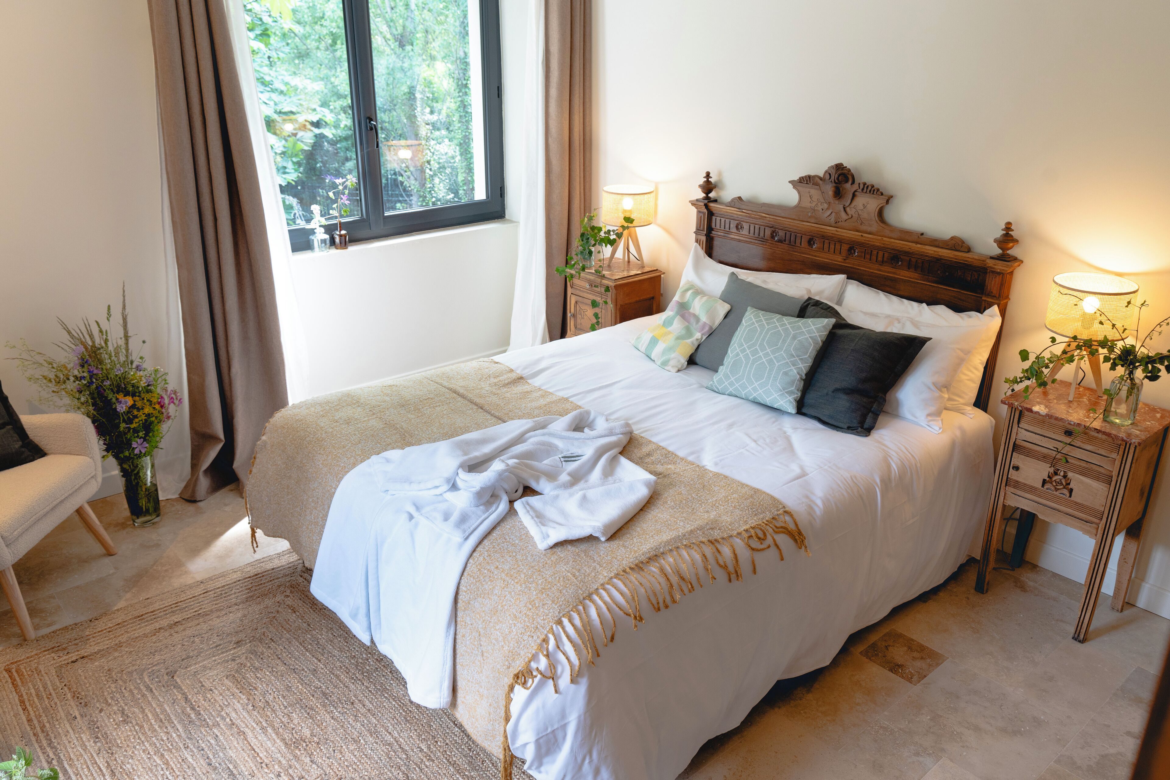 A neatly made bed with a wooden headboard, two bedside tables with lamps, a chair, and a window with curtains.