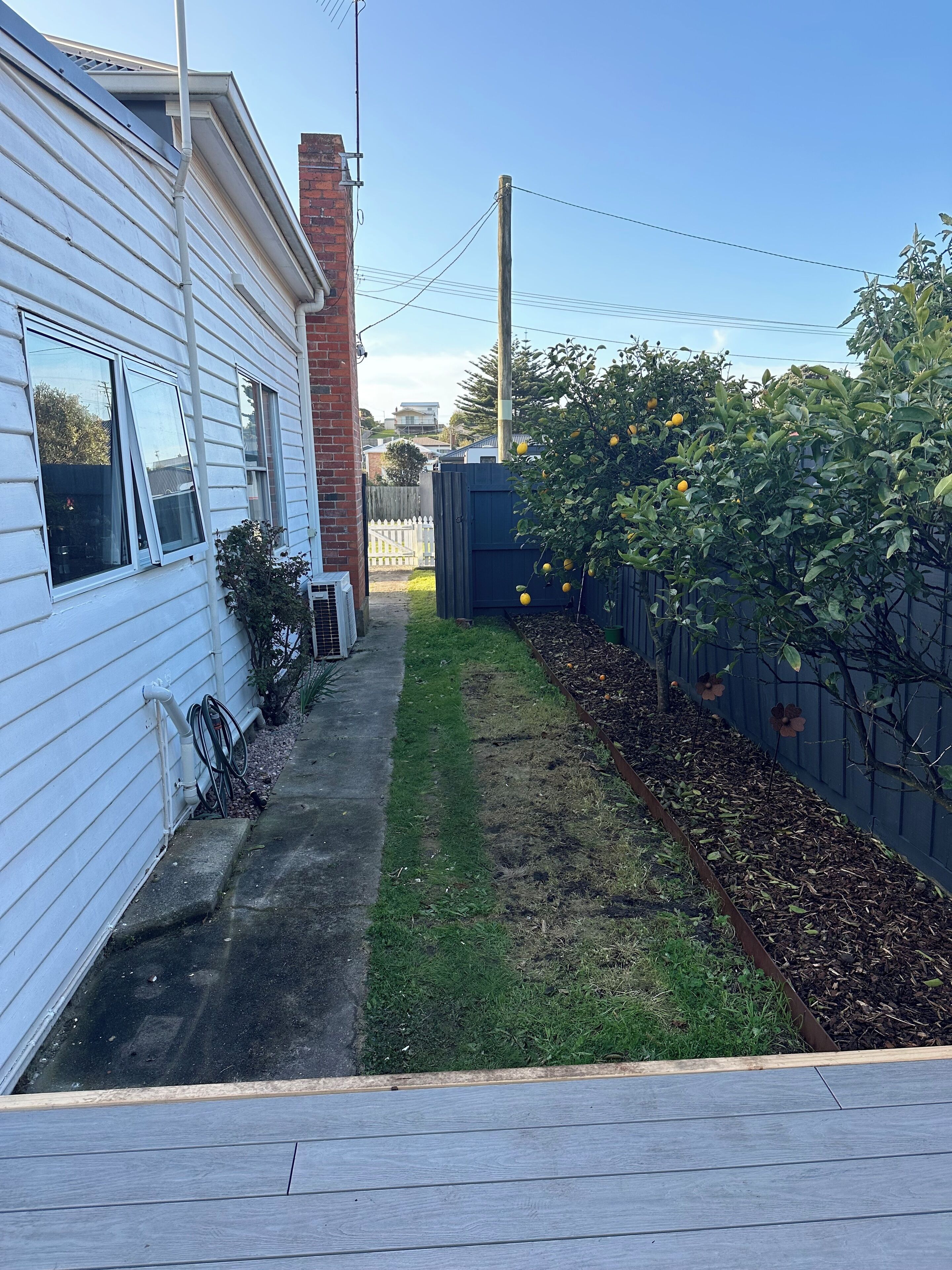 Un jardin à l’arrière d’une maison, avec une maison blanche, une terrasse en bois, un parterre fleuri et une clôture.