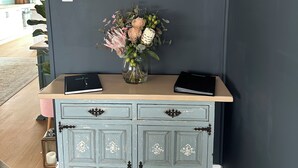Una parete blu scuro con un quadro floreale incorniciato, un vaso con fiori e un mobile azzurro con due quaderni neri sopra.