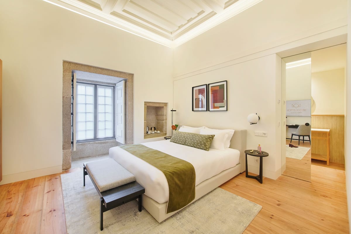 A bedroom with a bed, a bench, a small table, a lamp, and a desk with a chair.