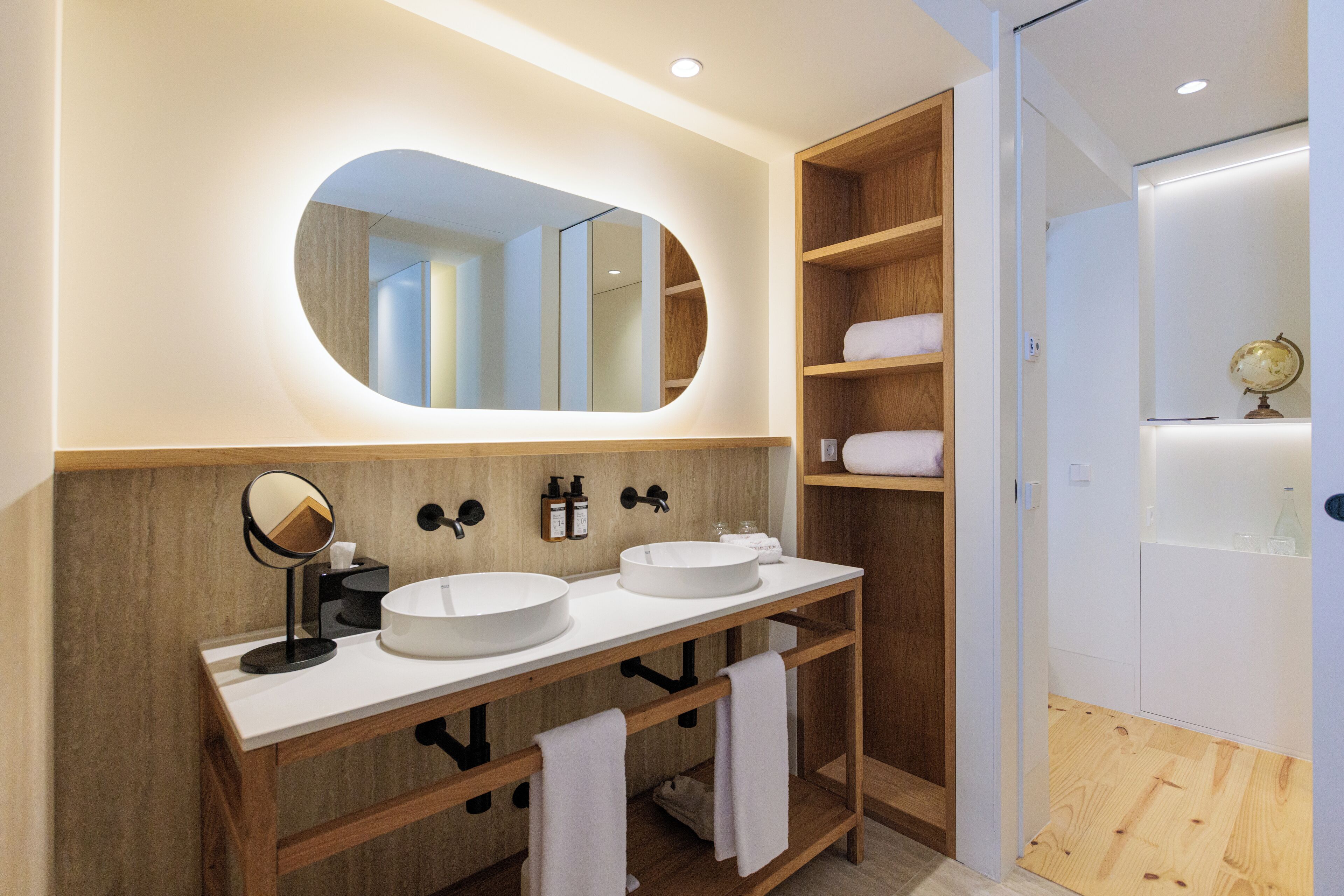 A modern bathroom with two sinks, a round mirror, wooden shelving, and a globe on a shelf.
