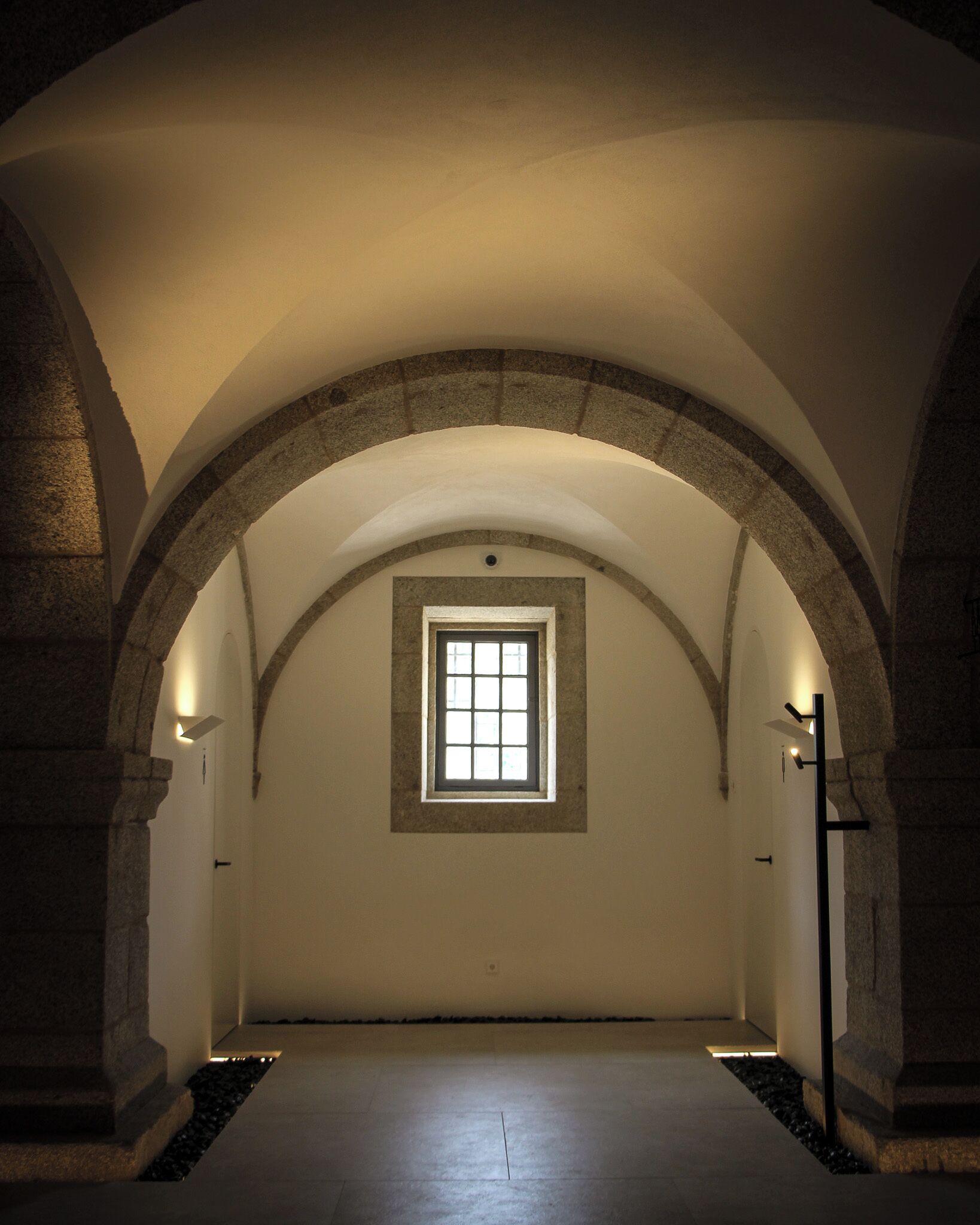 An arched corridor with a small window at the end, stone walls, and a tiled floor.