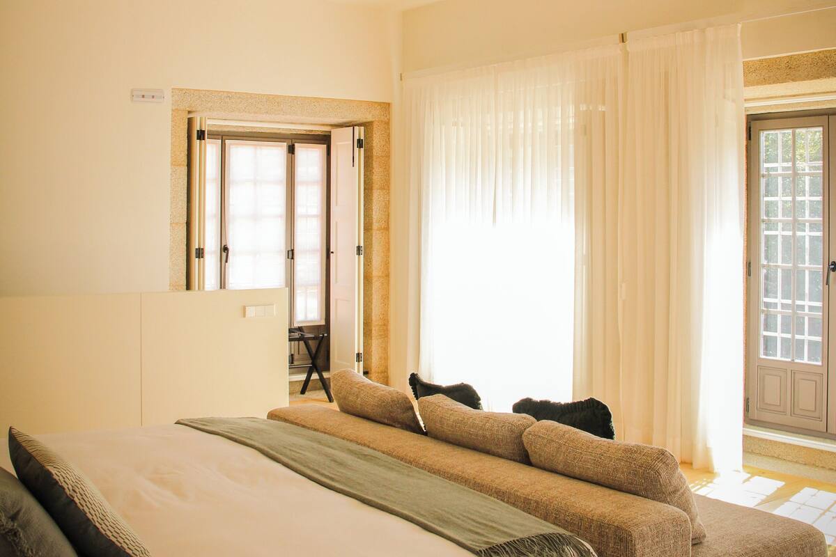 A bedroom with a bed, a sofa, a window with curtains, and a door.
