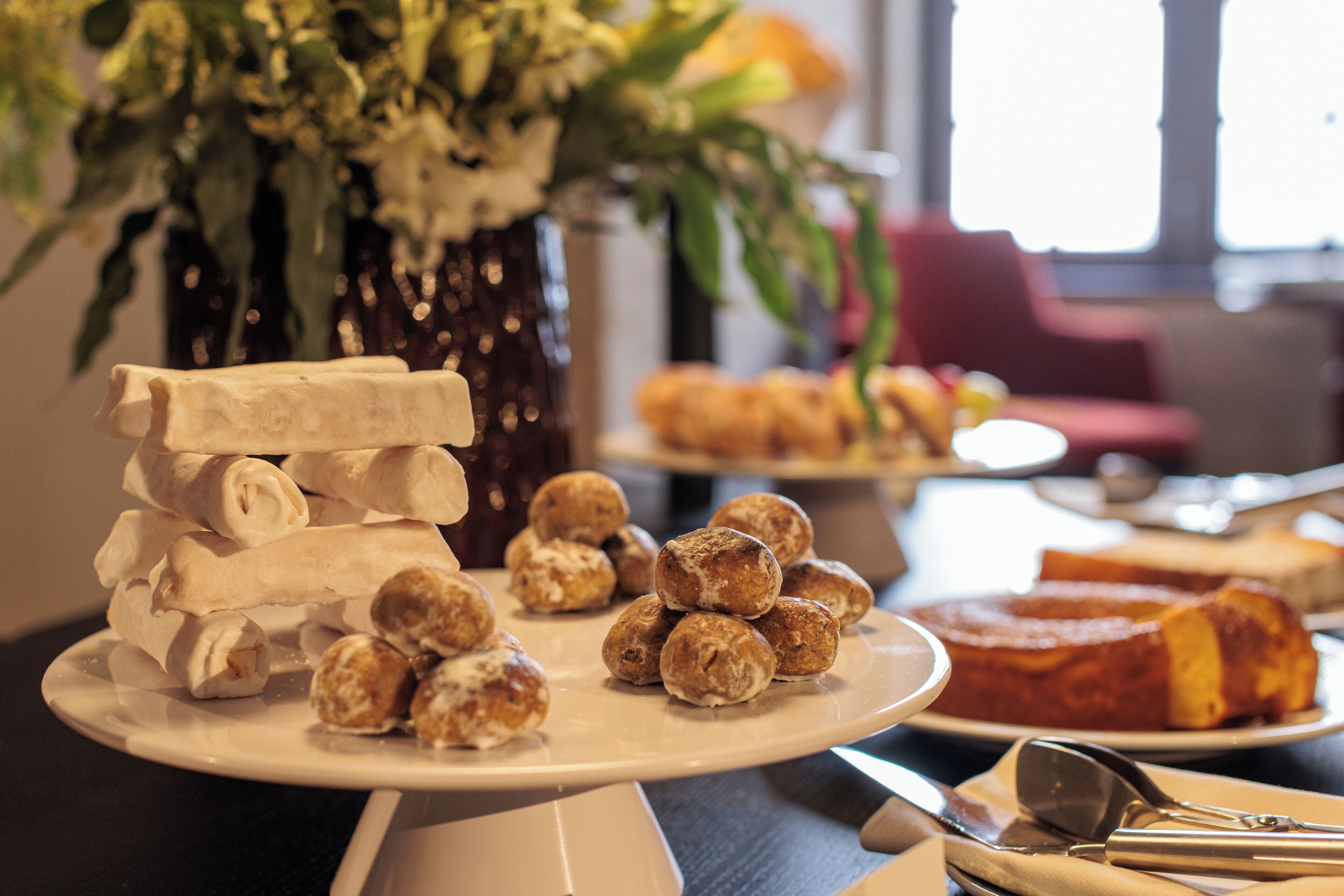 A dining table with various pastries and a cake, a vase with flowers, and a window in the background.