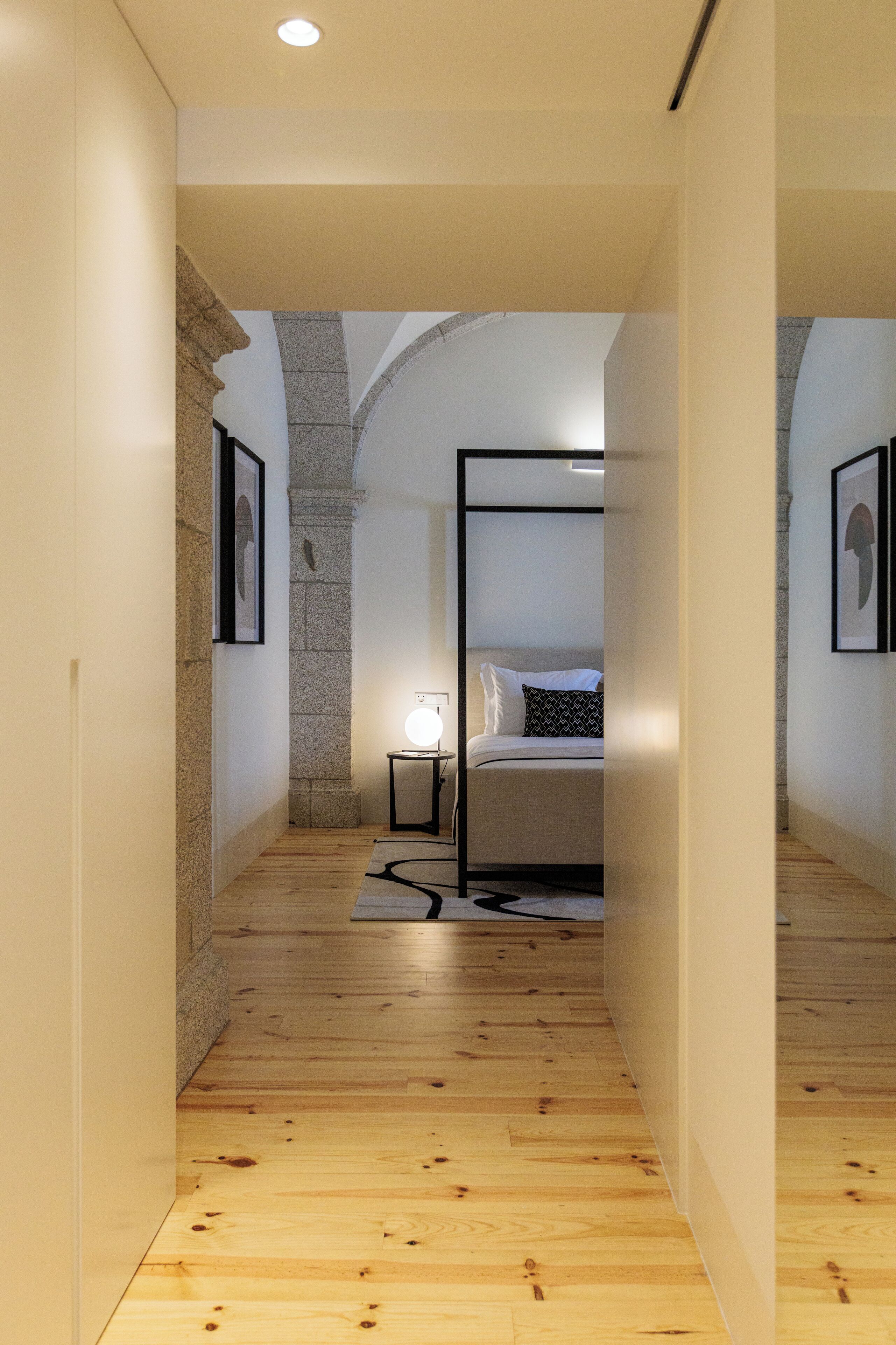 Un couloir étroit, doté d’un plancher en bois, mène à une chambre à coucher qui contient un lit, une table de chevet et un luminaire fixé au mur.