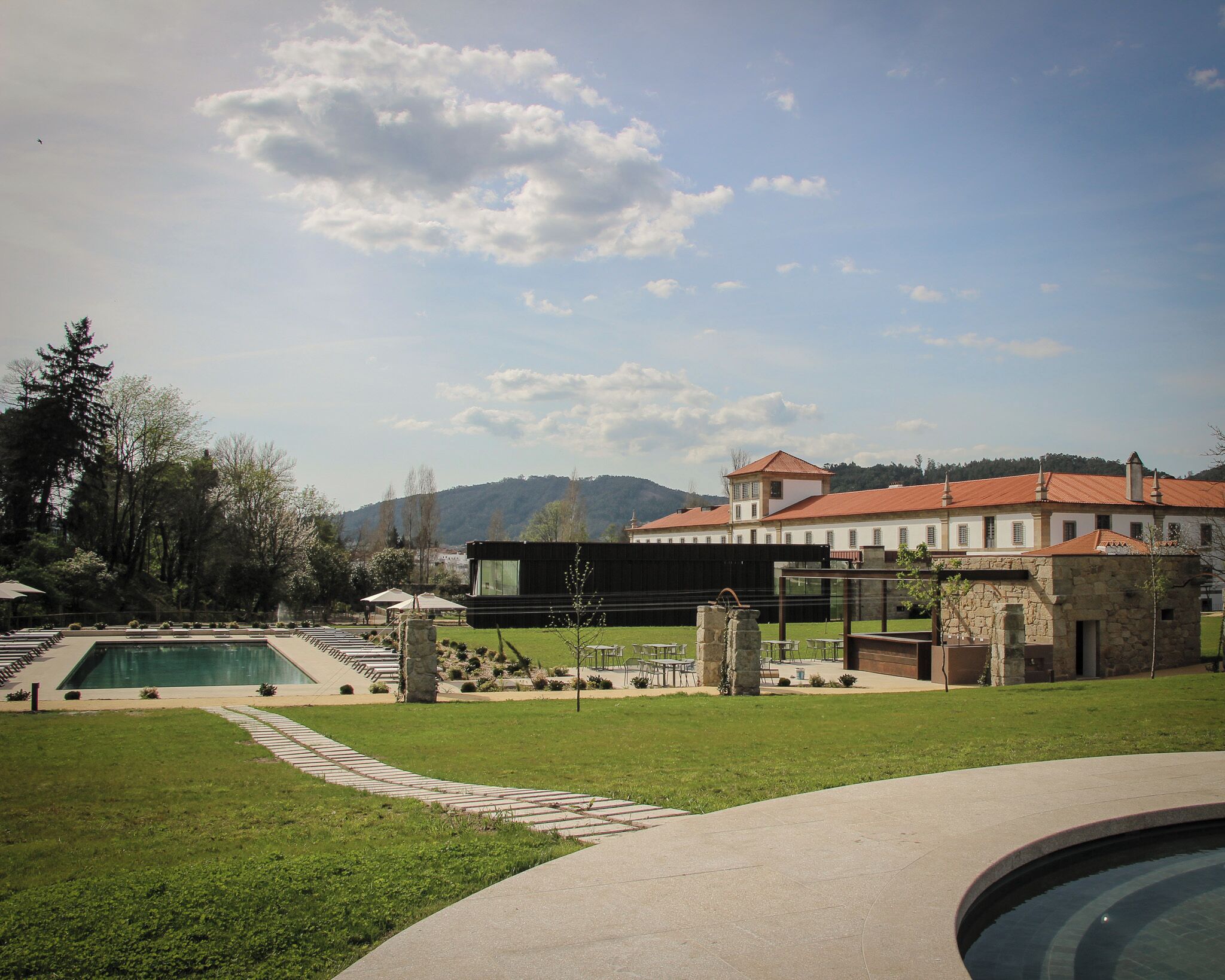 Área de piscina com um edifício moderno ao fundo, rodeada por vegetação e céu claro.