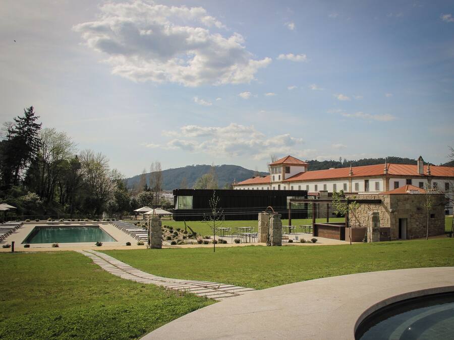 Área de piscina com um edifício moderno ao fundo, rodeada por vegetação e céu claro.