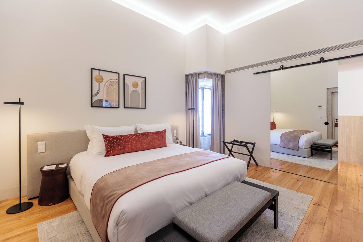 A modern hotel room with a large bed, a bedside table, a mirror, and a view of another room.