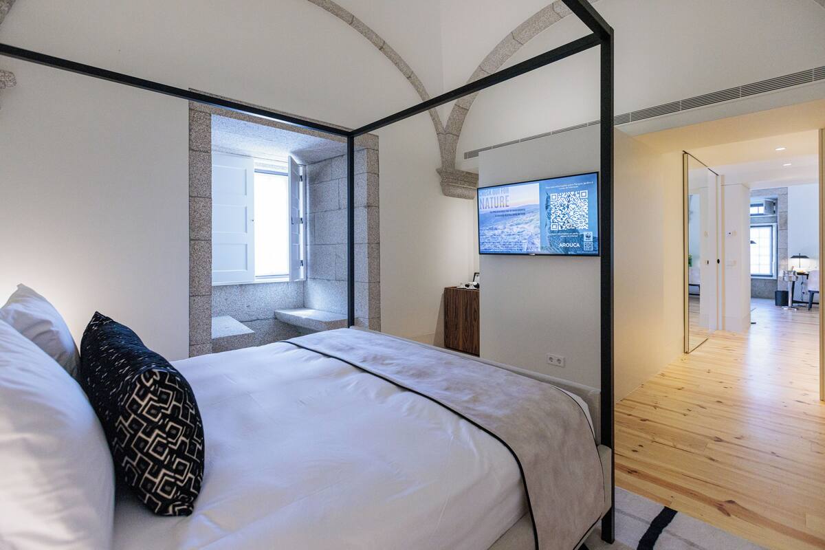 A modern bedroom with a four-poster bed, a stone accent wall, a mounted TV, and a wooden floor.