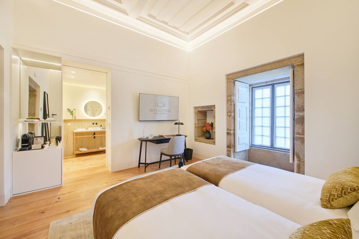 A modern hotel room with a wooden floor, two beds with brown headboards, a desk with a chair, a small bathroom, and a large window.
