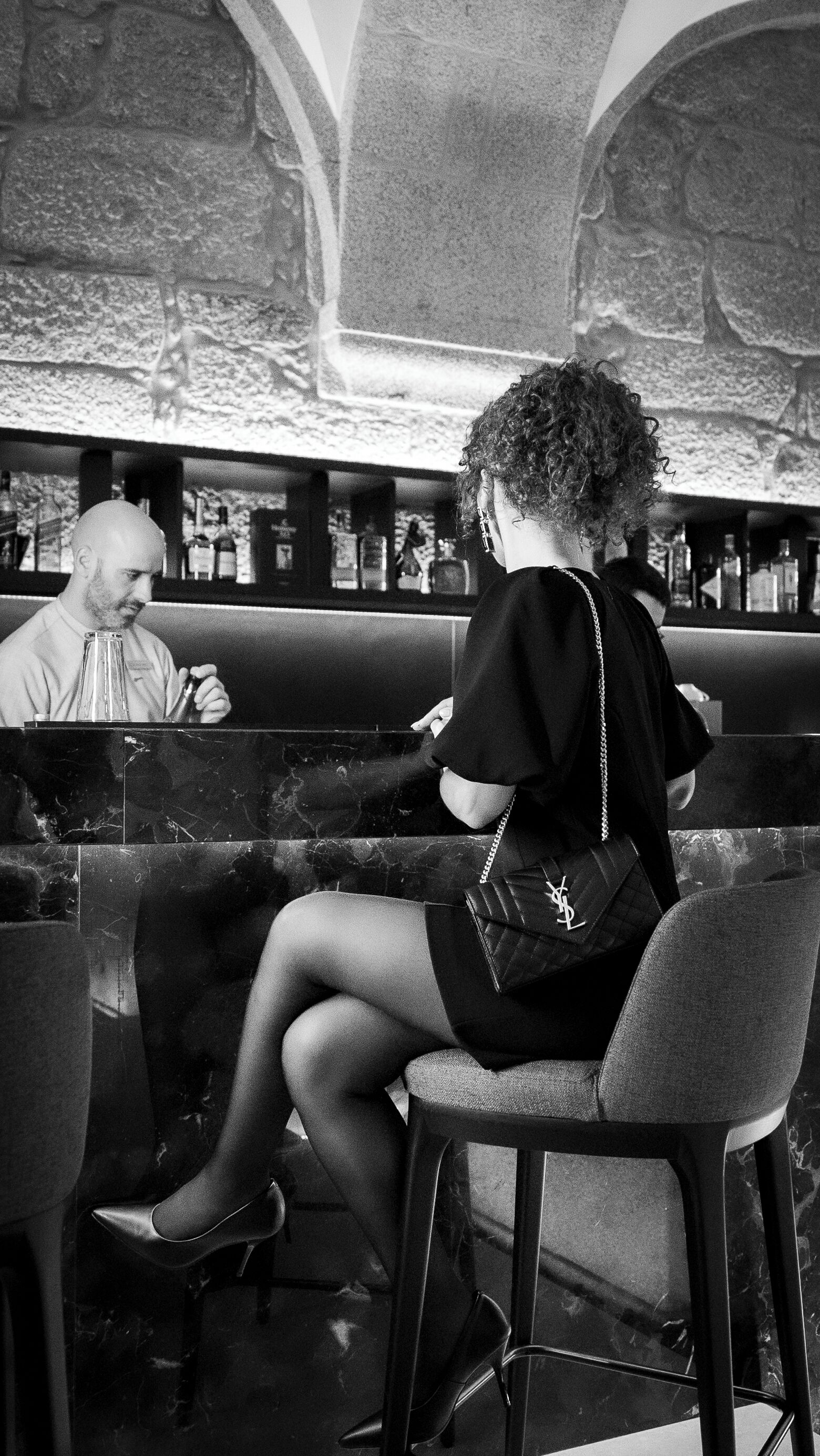 A person seated at a bar with a drink, a bar counter, and shelves with bottles in the background.