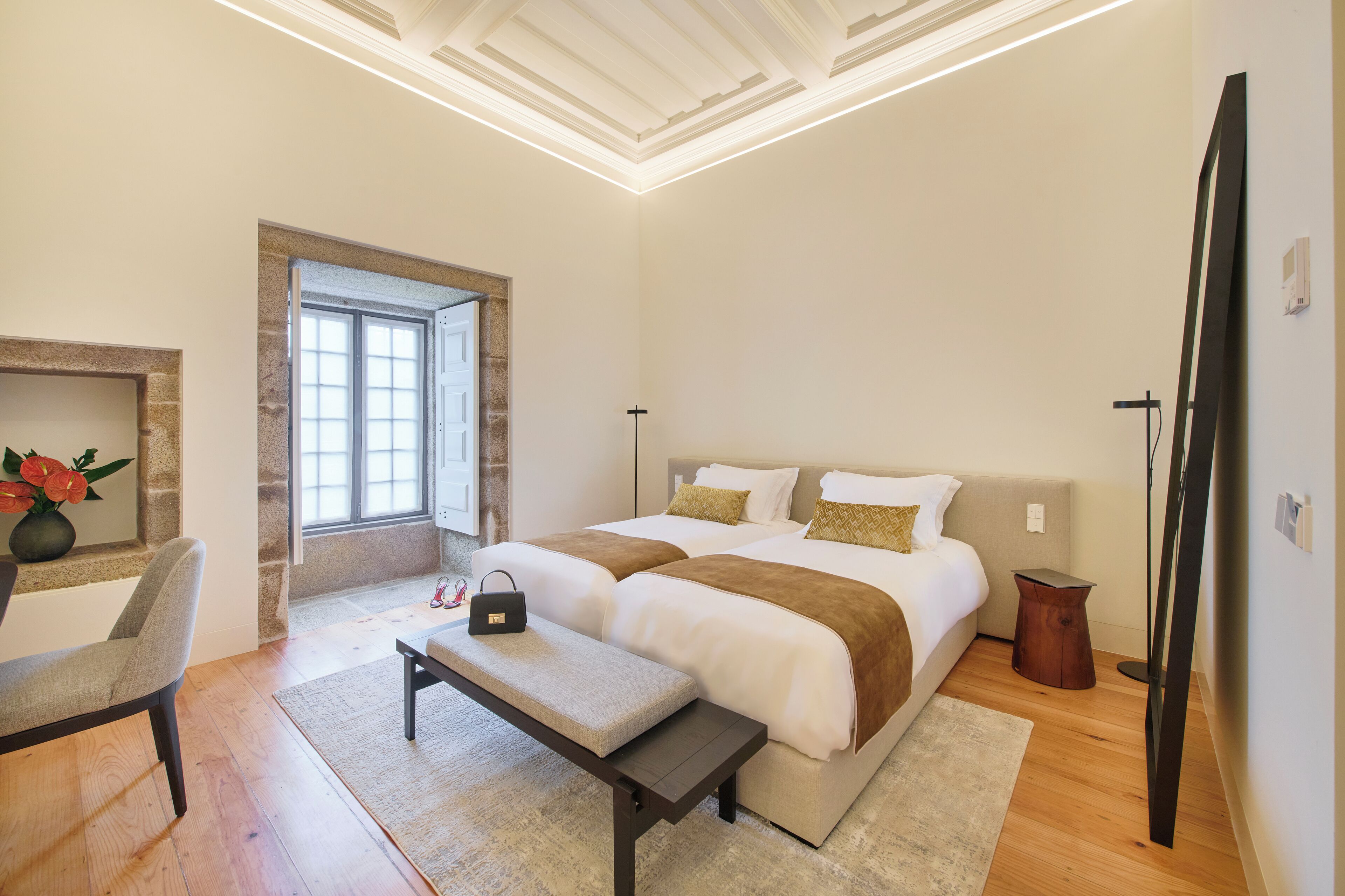 A bedroom with a large bed, a bench, a chair, a bedside table, a lamp, and a window.