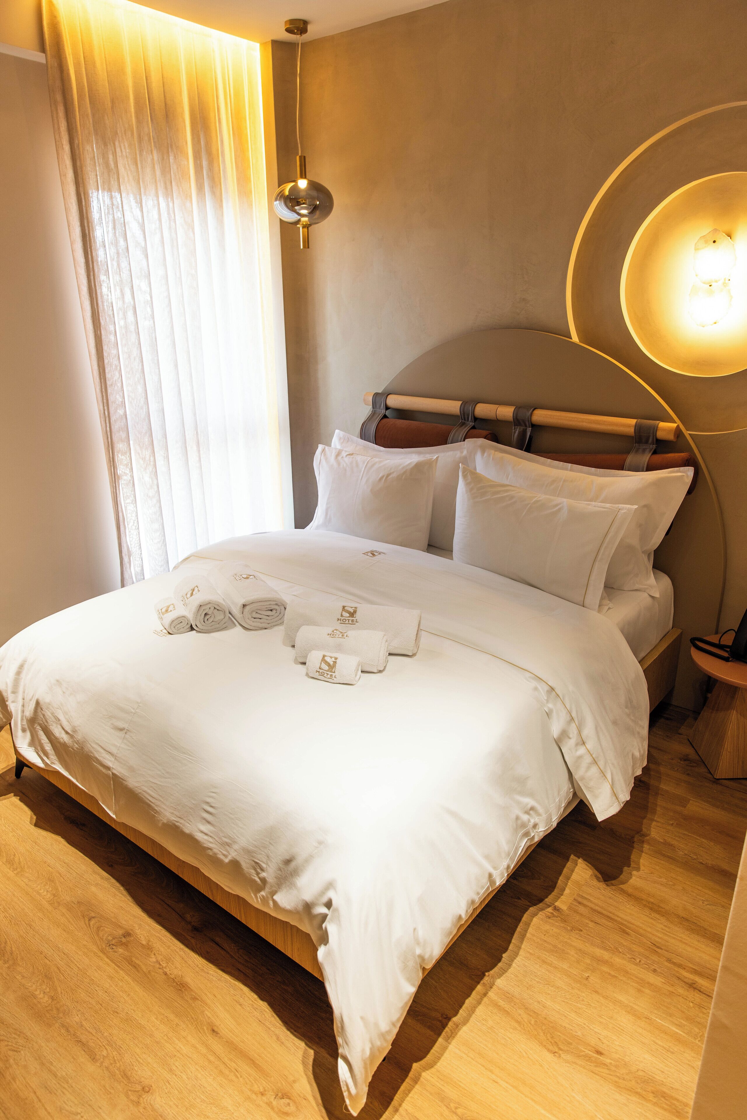 A neatly made bed with white linens and towels, a wooden headboard, a bedside lamp, and a wooden floor.