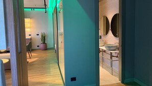 Ein modernes Hotelzimmer mit Holzboden, einer Sitzecke mit Stuhl und Tisch, einer Zimmerpflanze und einem Badezimmer mit großem Spiegel und Waschbecken.