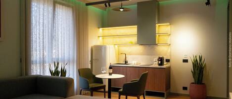 A modern living room with a sofa, a dining table, and chairs. There is a kitchen area with a refrigerator and a sink.