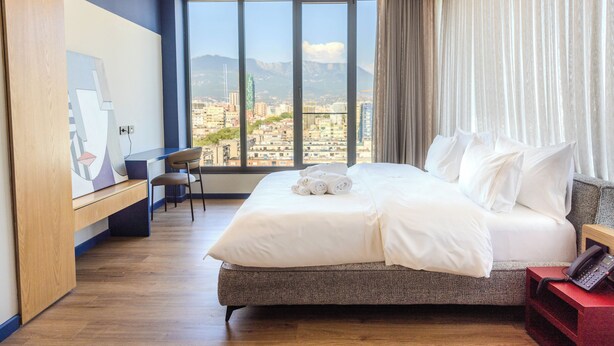 Ein modernes Hotelzimmer mit einem großen Bett, einem Schreibtisch und Blick auf eine Stadtlandschaft durch große Fenster.
