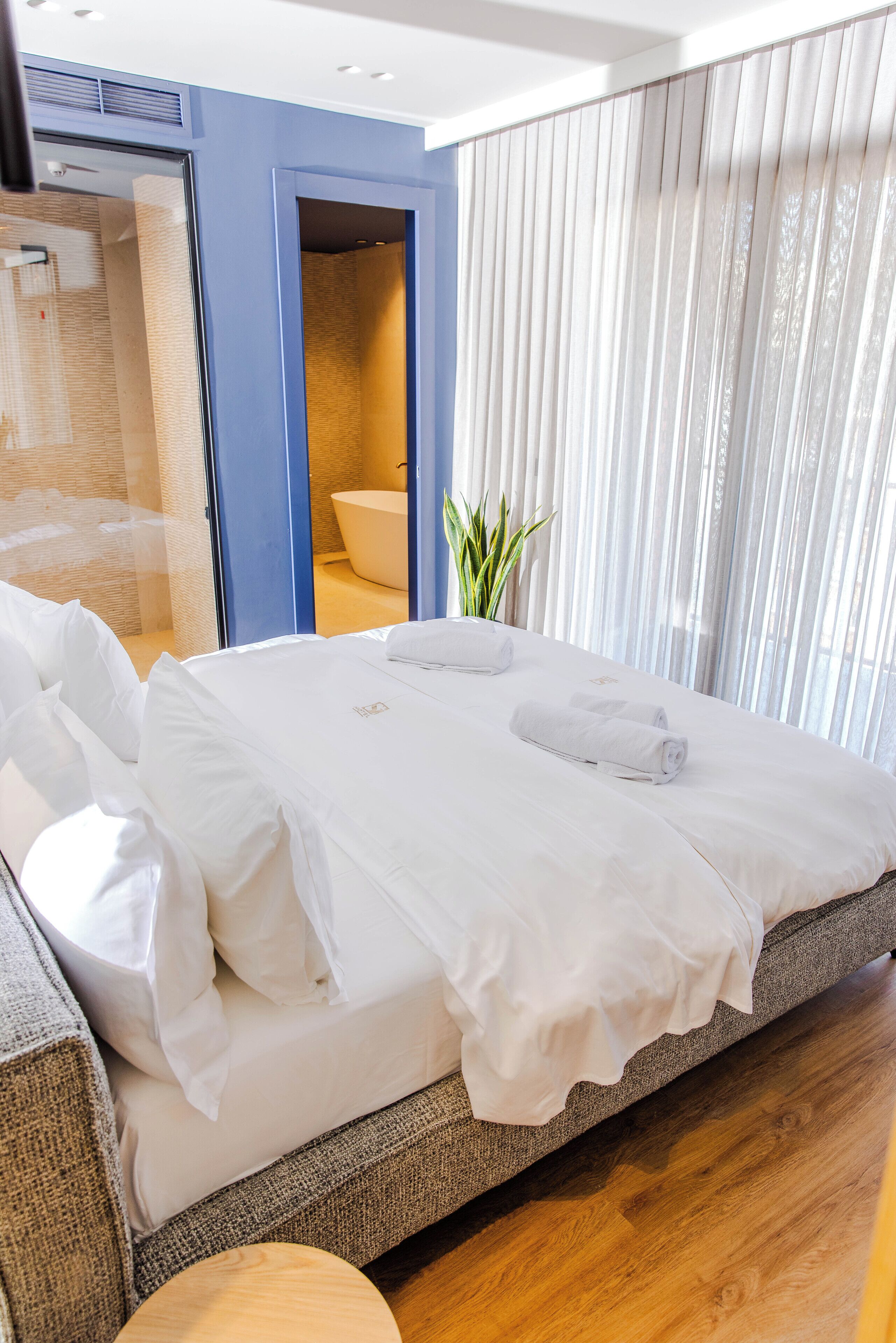 A modern hotel room with a large bed, a bathroom visible through an open door, and a potted plant.
