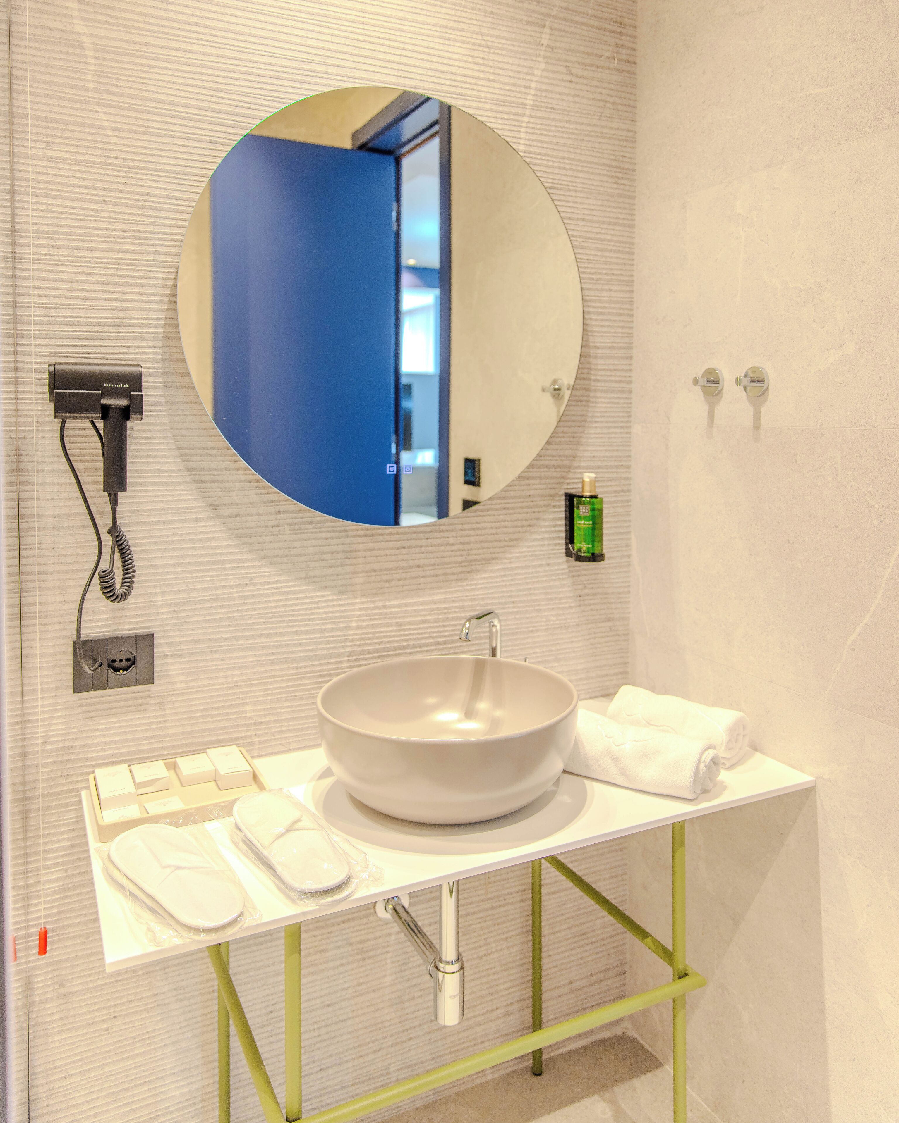 Een badkamer met een ronde spiegel, een witte wastafel, een handdoekhouder, een haardroger en een groene fles.