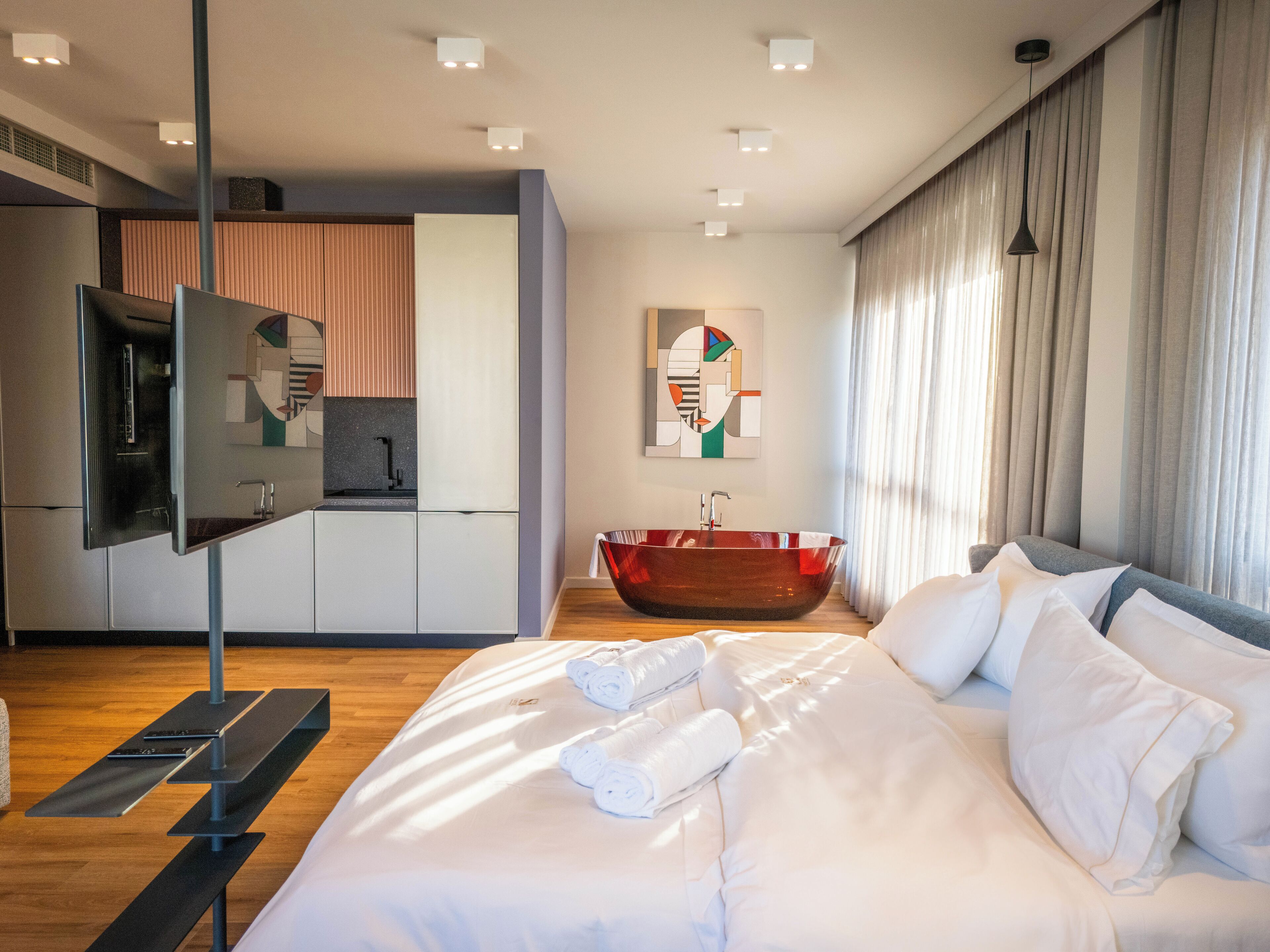 A modern hotel room with a large bed, a built-in desk, a red glass bathtub, and a contemporary art piece on the wall.