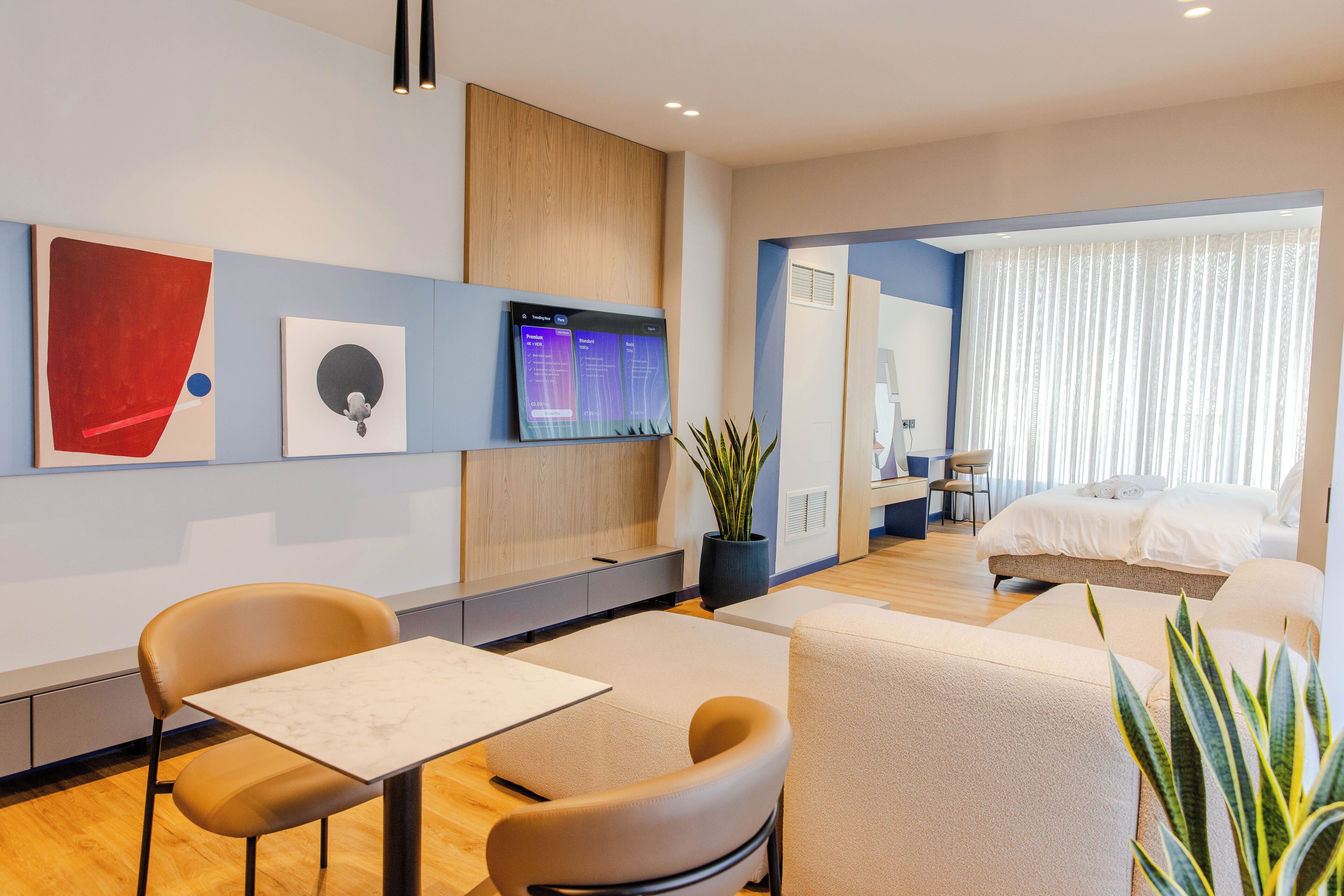 Habitació d'hotel moderna amb un llit, zona de seients amb taula i cadires, una finestra gran amb cortines i obres d'art a les parets.