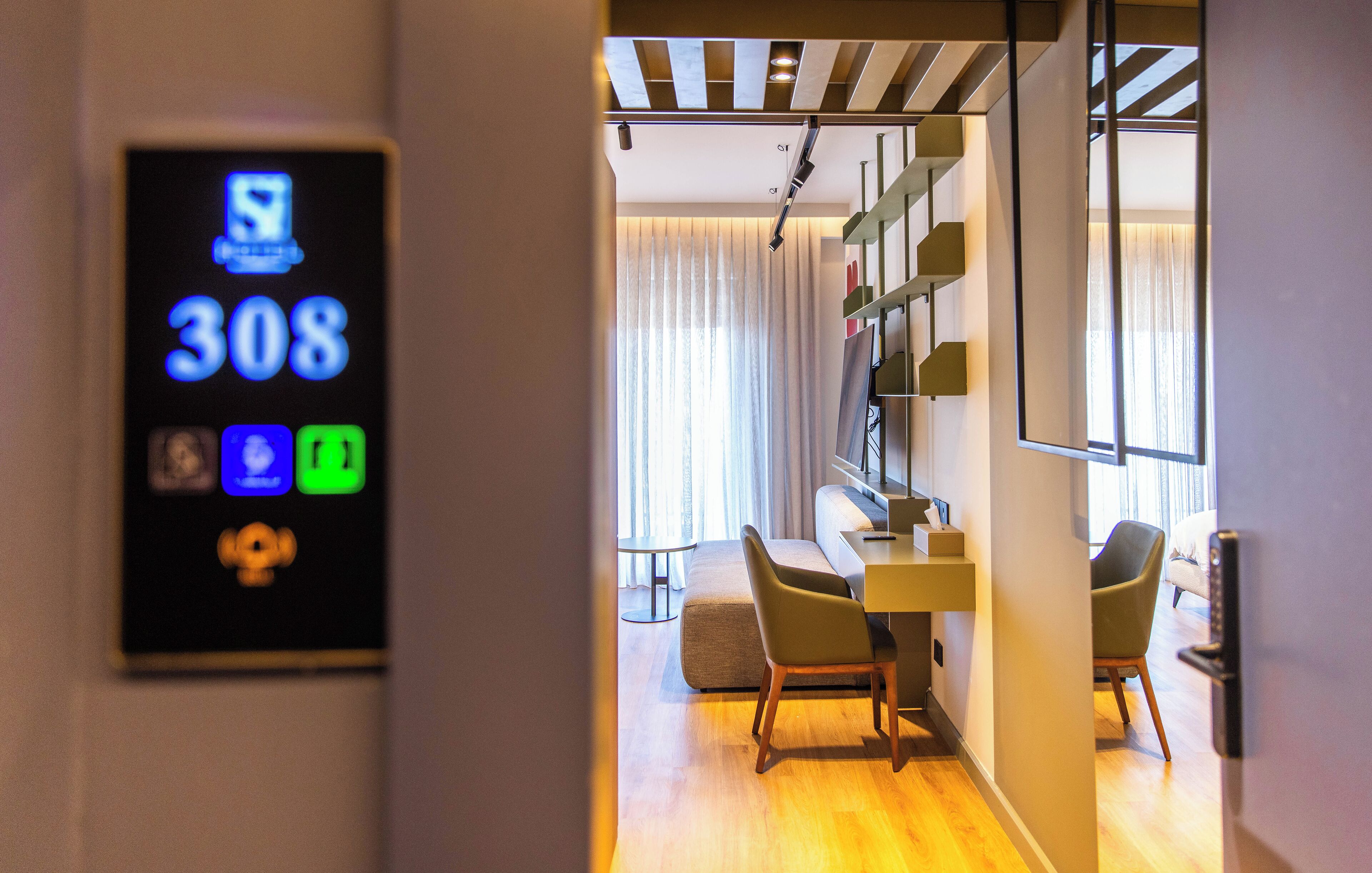 Una habitación de hotel moderna con una pantalla digital que muestra el número de habitación 308, una zona de comedor con una mesa y sillas, y un ventanal con cortinas.